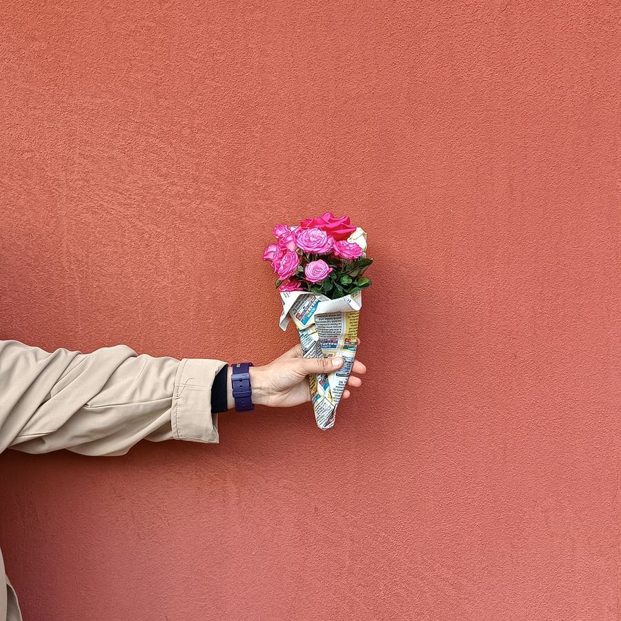 A hand holding a small bouquet of roses wrapped in paper against a red wall