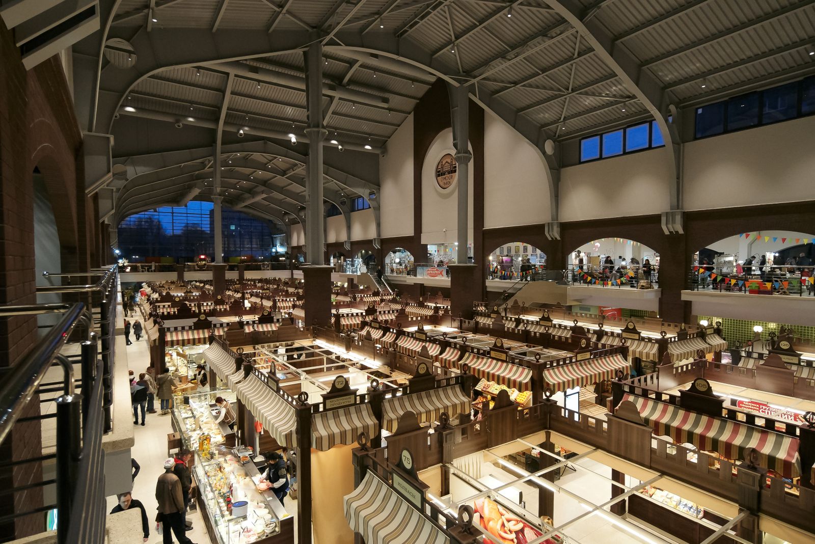 A large market hall under an arched roof, seen from above, with rows of stalls and people walking between them