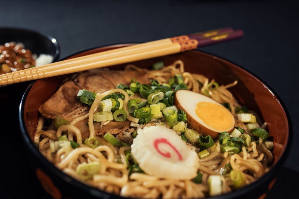 A bowl of ramen with a boiled egg, spring onions and chopsticks on a dark table