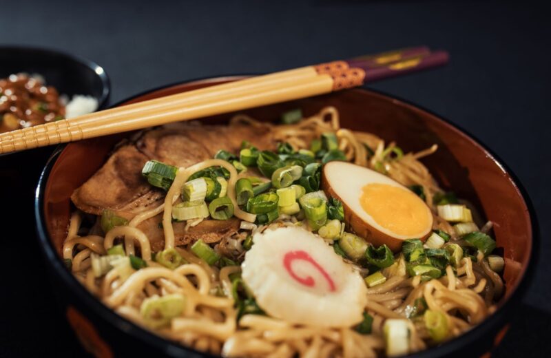 A bowl of ramen with a boiled egg, spring onions and chopsticks on a dark table