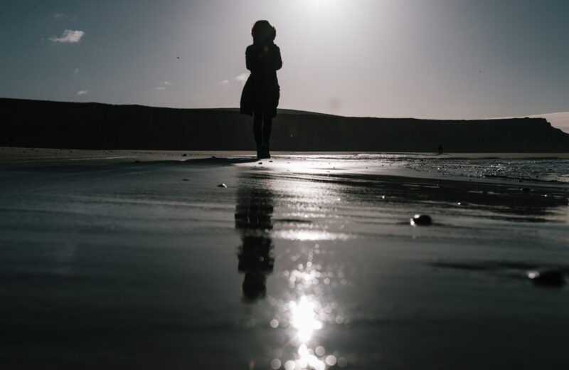 A person walking along the shore at sunset, reflected in the wet sand