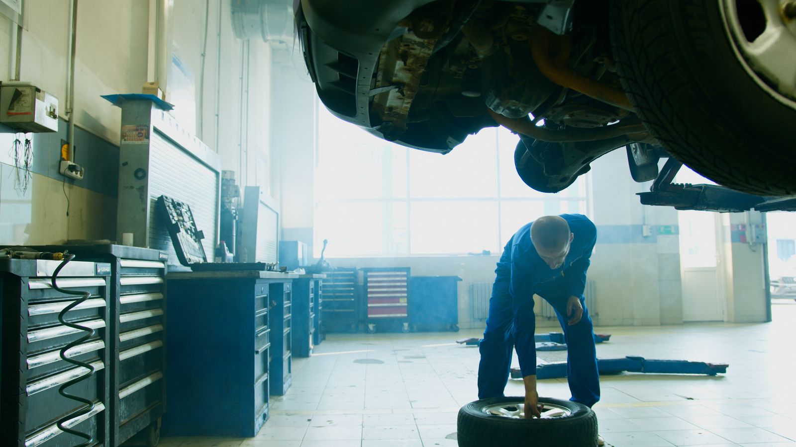 A mechanic rolling a wheel and tyre under a car raised on a lift