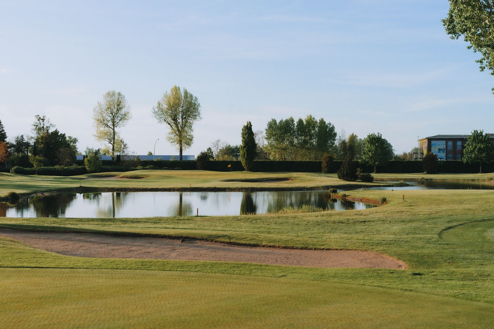 A green beside a still pond on a summer day, trees and a clubhouse roof behind