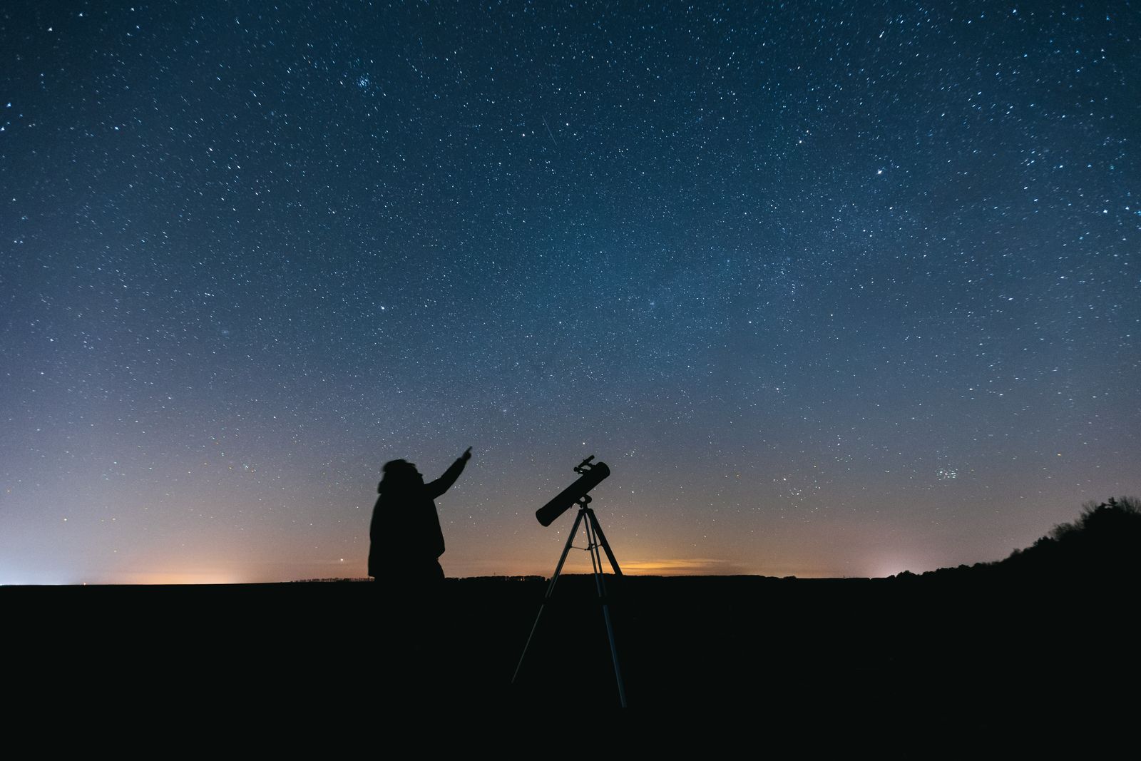 A person in silhouette pointing at the night sky beside a small telescope on a ridge