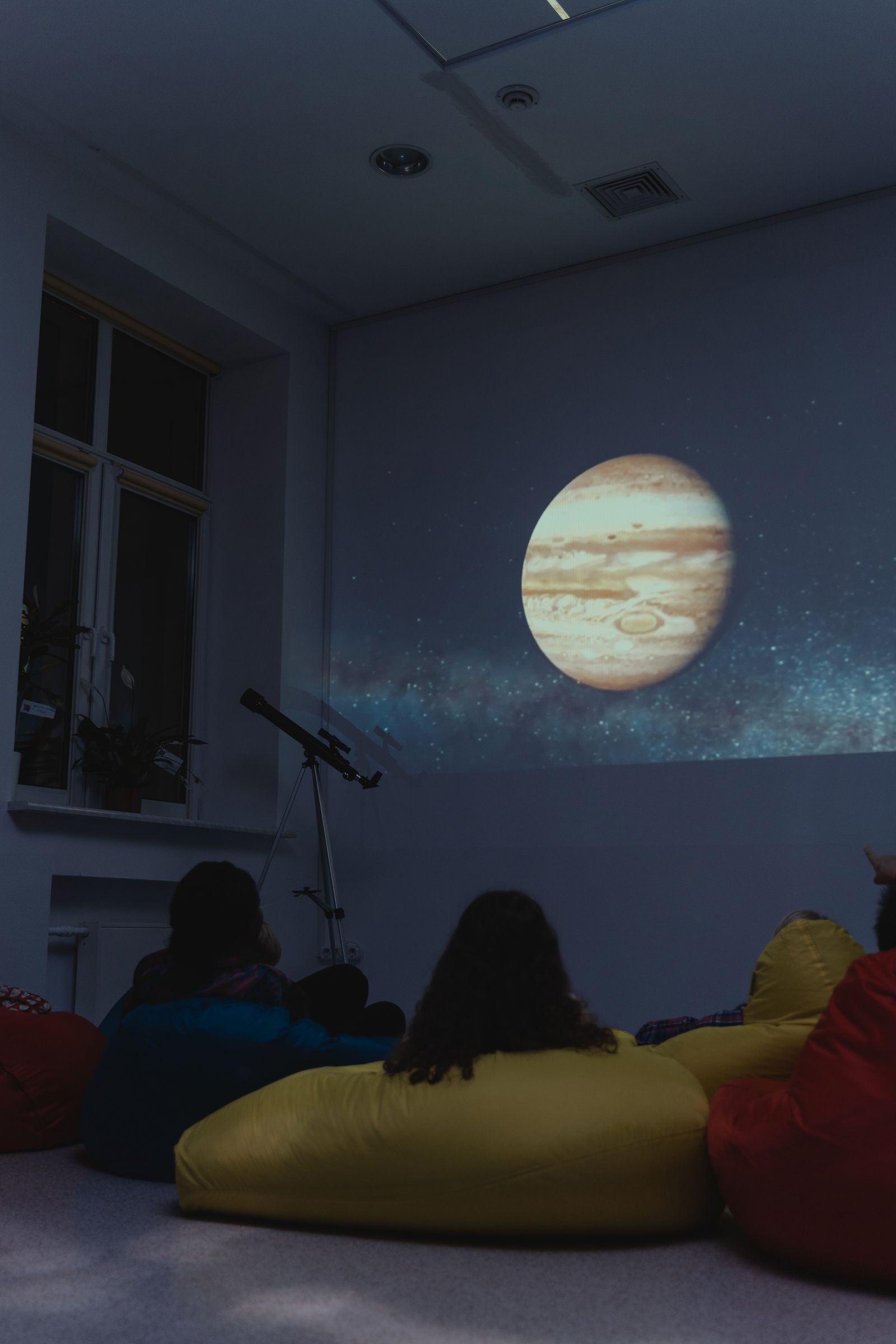 A class seen from behind on floor cushions watching Jupiter projected on a darkened wall