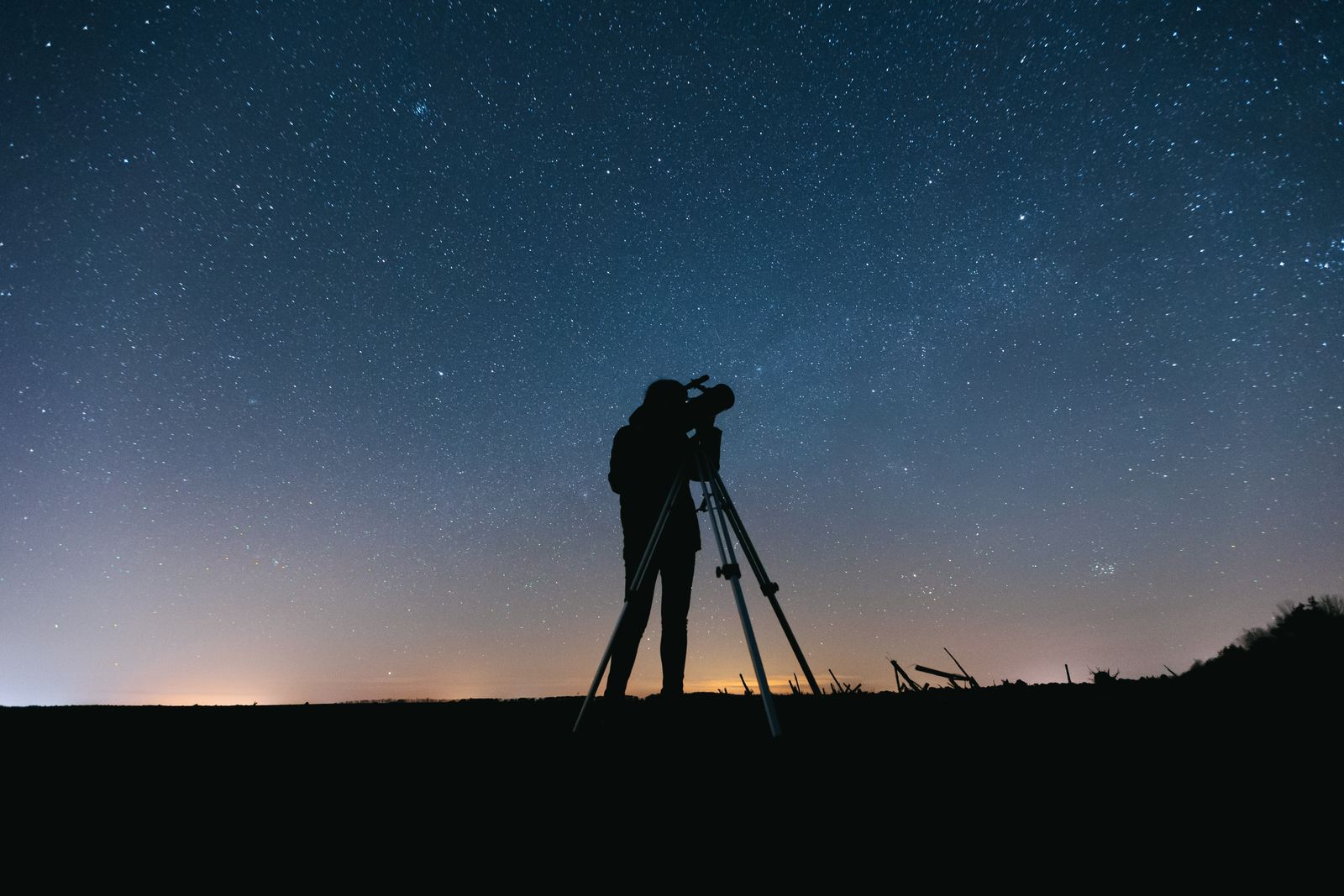 A person in silhouette looking through a telescope on a tripod under a sky full of stars