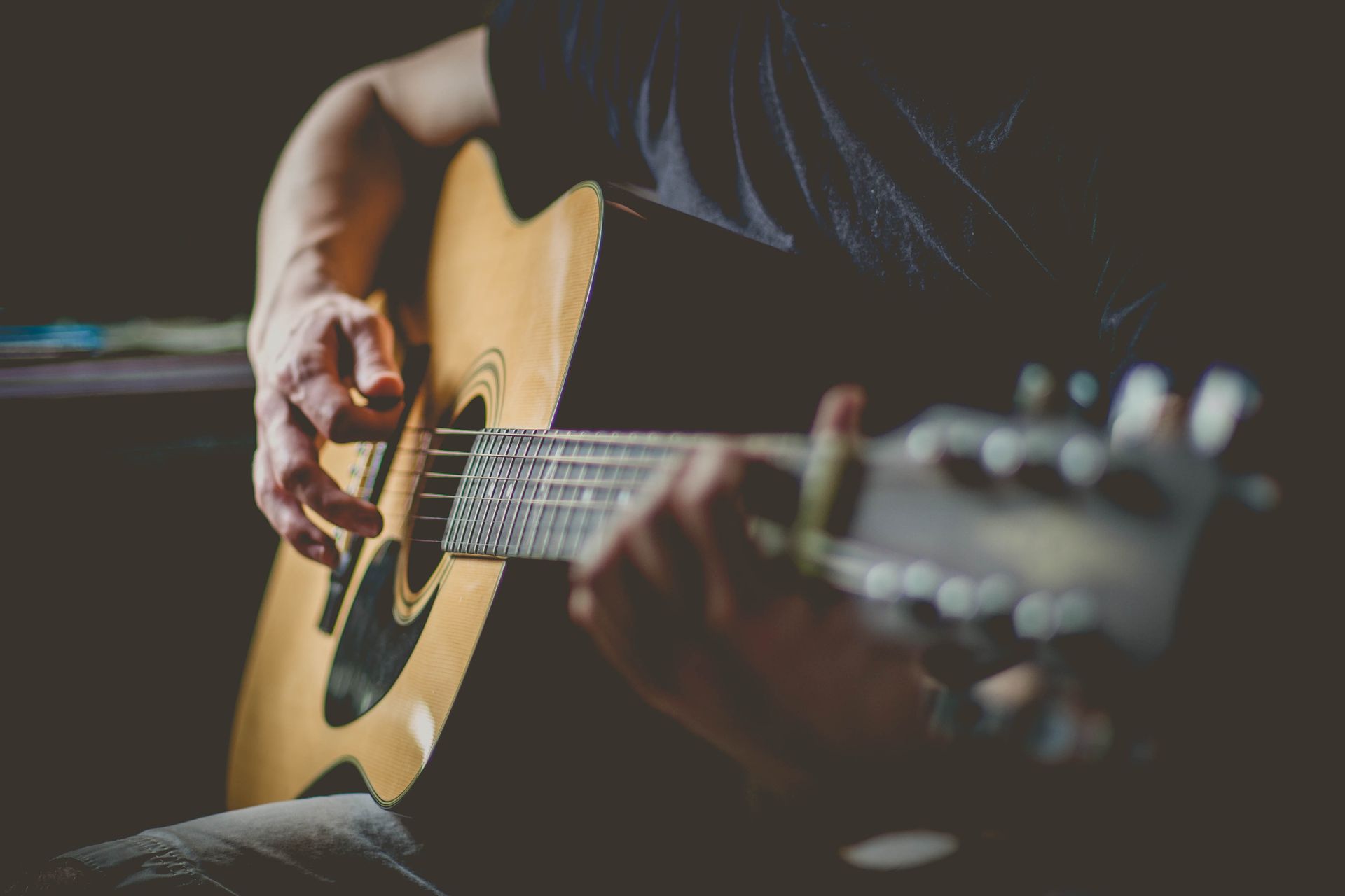 Hands playing a natural-finish acoustic guitar in a dark room, fingers on the fretboard