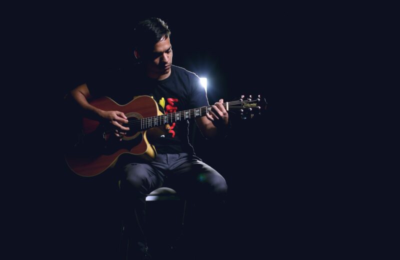 A guitarist playing an acoustic guitar on a dim stage under a single light