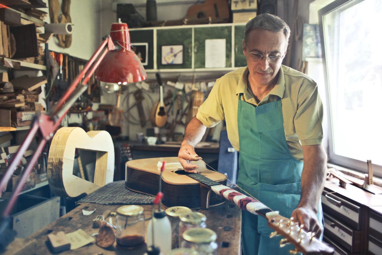 A luthier in an apron working on the neck of an acoustic guitar at a cluttered workbench