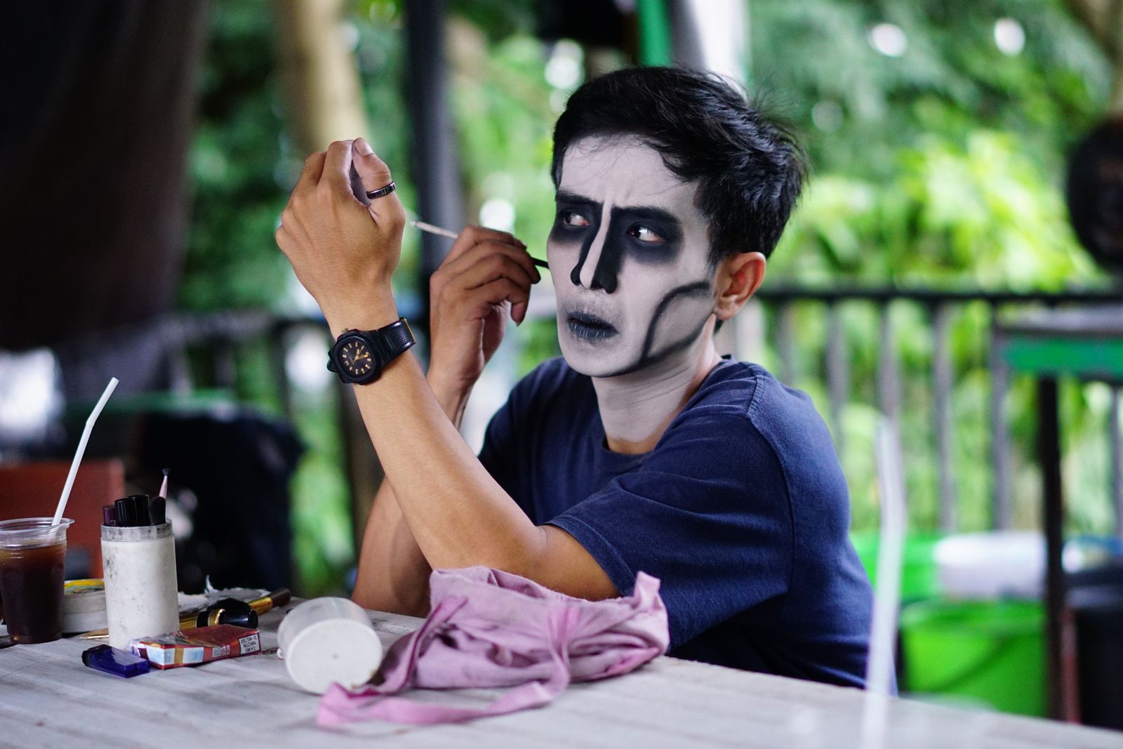 A man painting a dark skull design on his own face at an outdoor table with brushes and paint pots