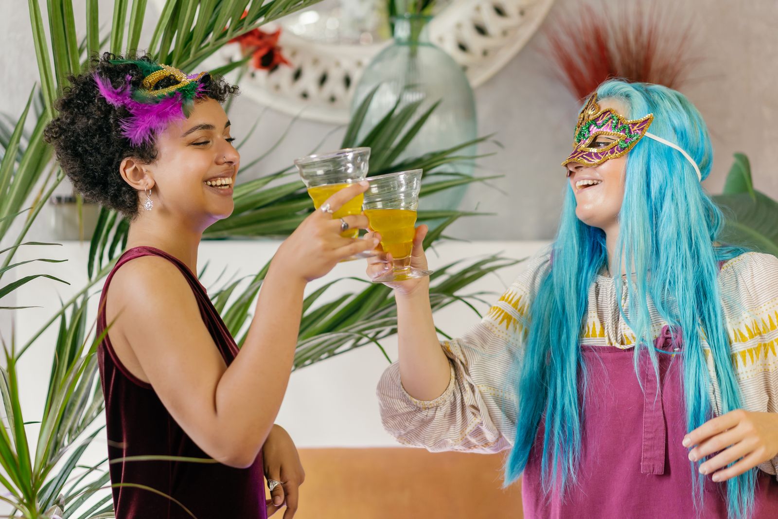 Two women in masquerade masks and bright wigs raising drinks at a themed party