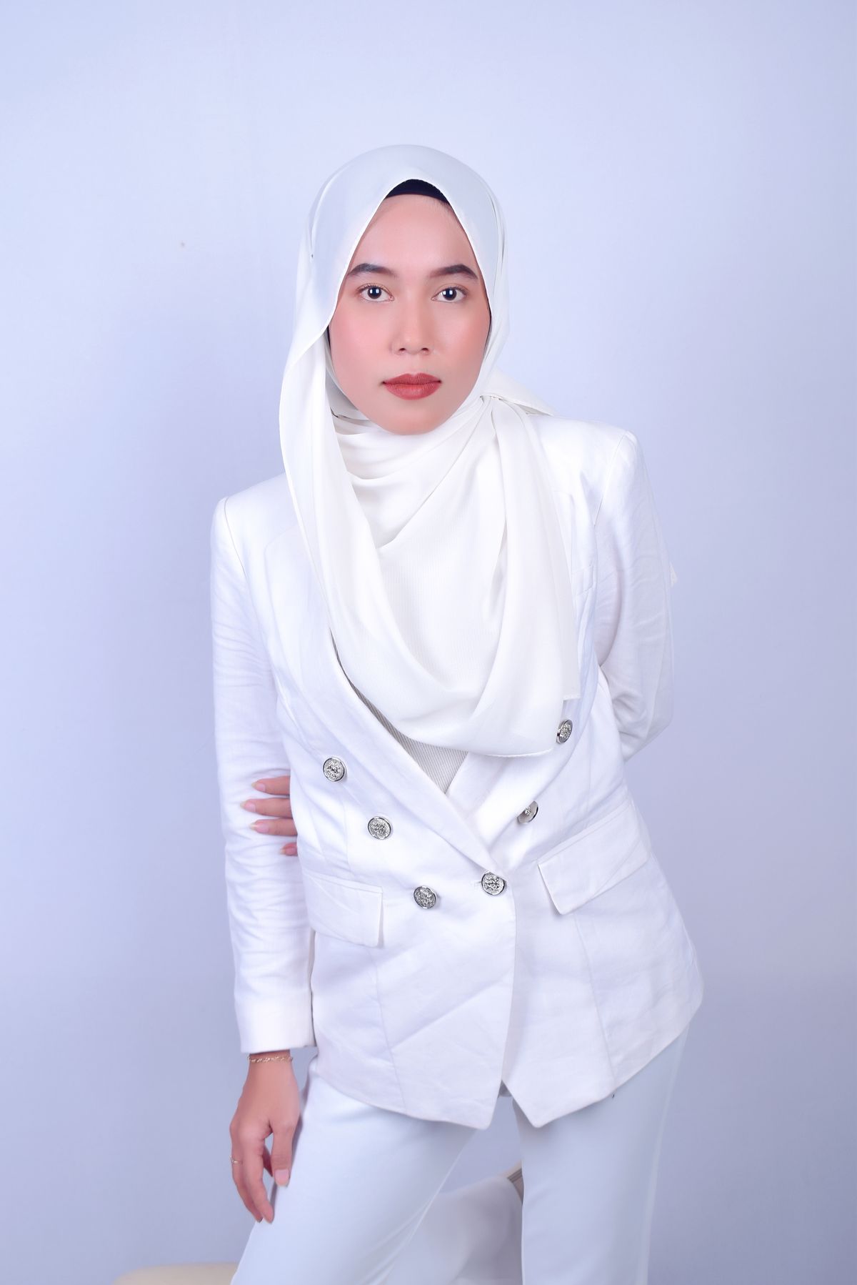 A woman in an ivory hijab draped over a white double-breasted suit, against a pale lilac wall