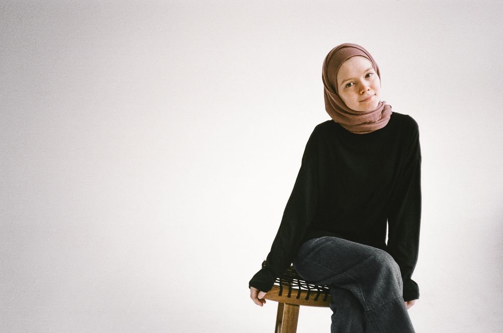 A woman in a taupe jersey hijab sitting on a wooden stool in a white studio