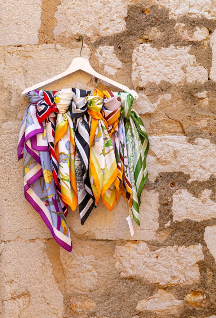 Printed silk square scarves in purple, orange and white hanging from a rail against a stone wall
