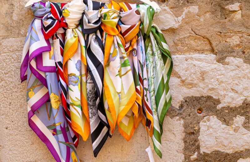 Printed silk square scarves in purple, orange and white hanging from a rail against a stone wall