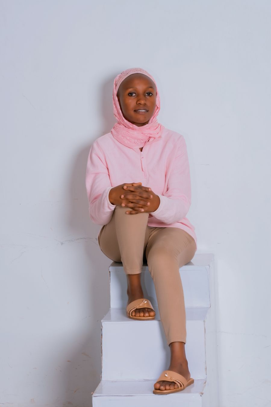 A woman in a blush hijab and pink sweatshirt sitting on a white block in a pale studio
