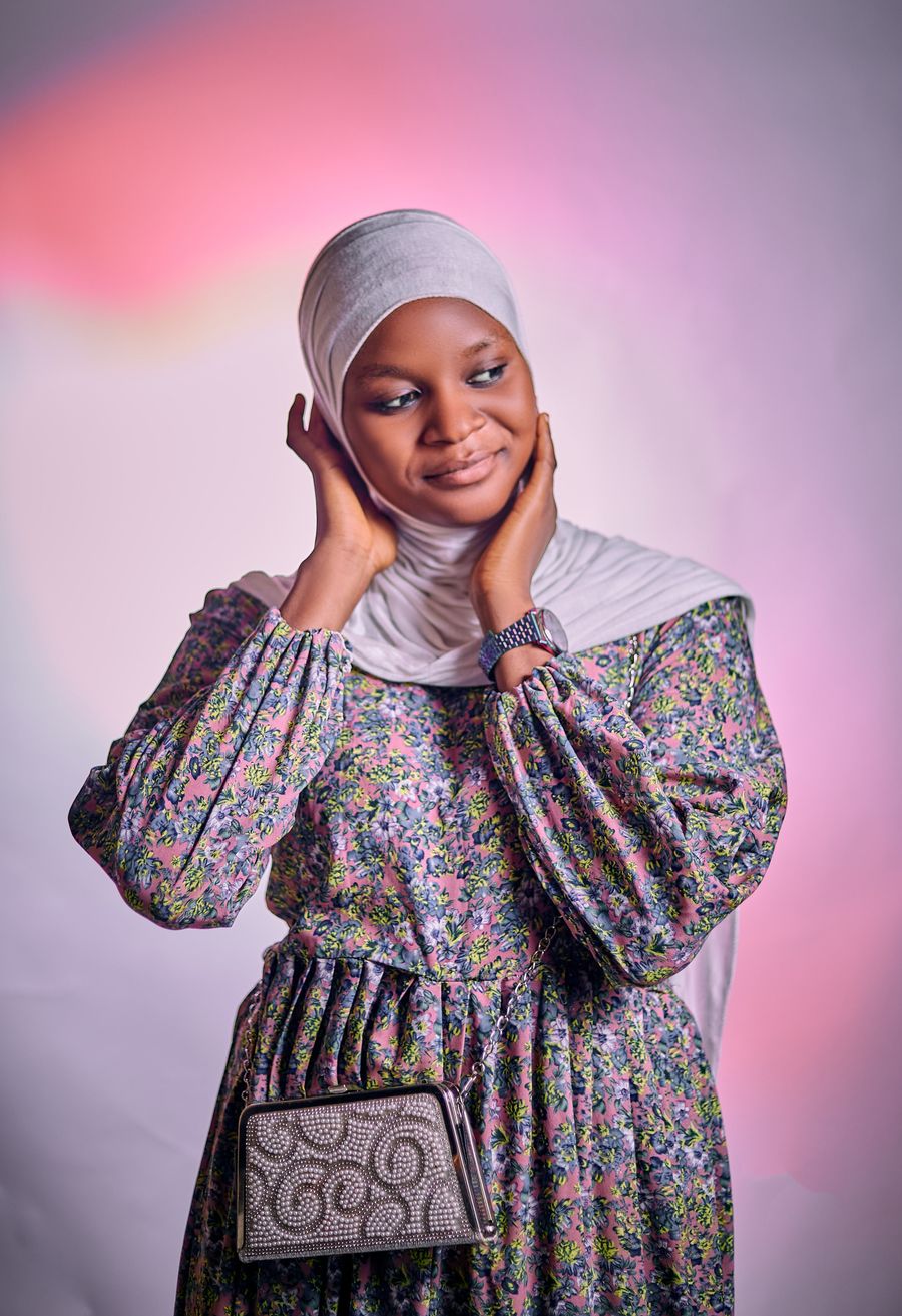 A woman in a grey hijab and a floral dress against a pink backdrop, hands at her face