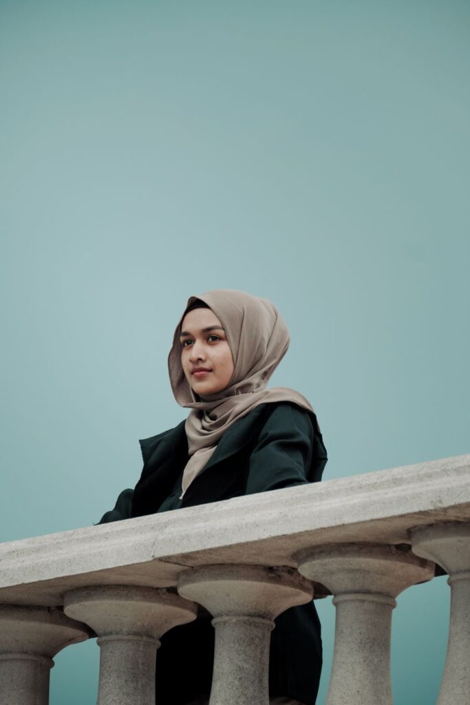 A woman in a sand-coloured hijab and black coat by a railing against a pale blue wall