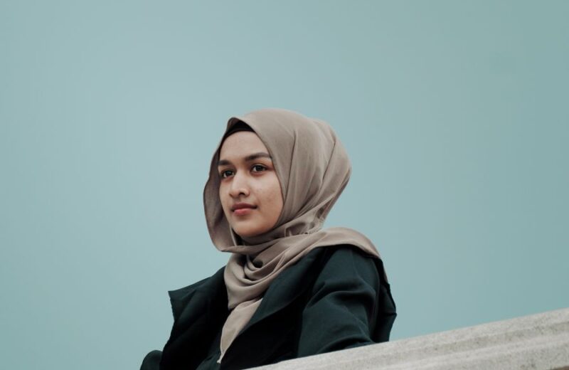 A woman in a sand-coloured hijab and black coat by a railing against a pale blue wall