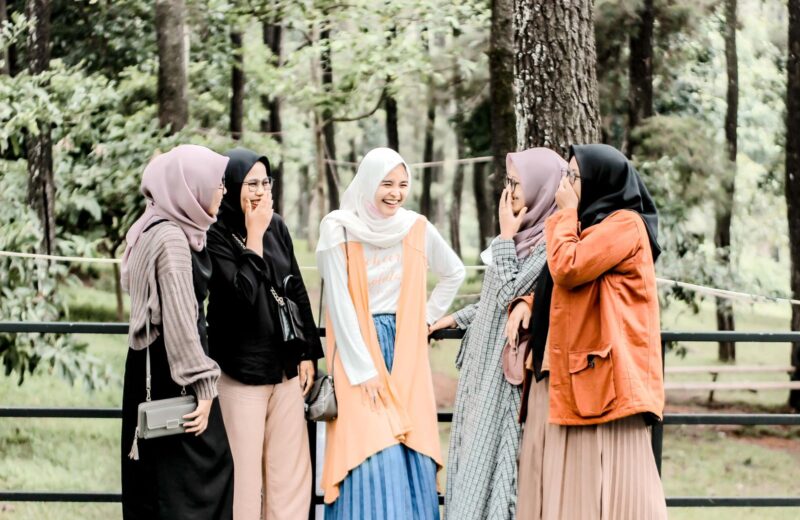Five women friends in hijabs and long skirts laughing together among trees