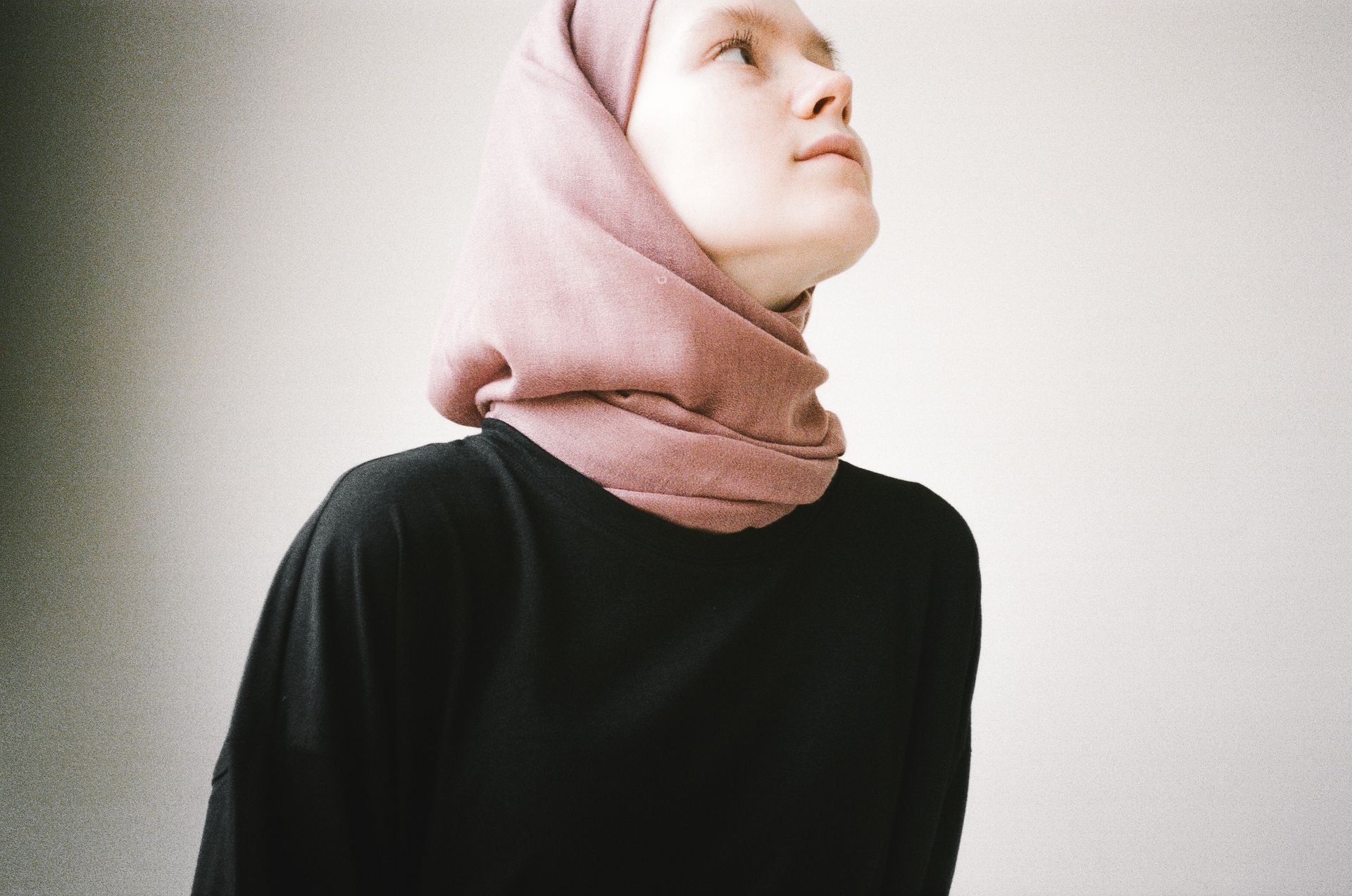 A woman in a blush pink hijab and black top looking up in a white studio