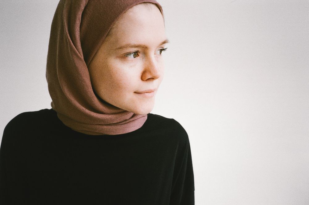 A woman in a soft brown hijab and black top in a bright white studio, looking to the side