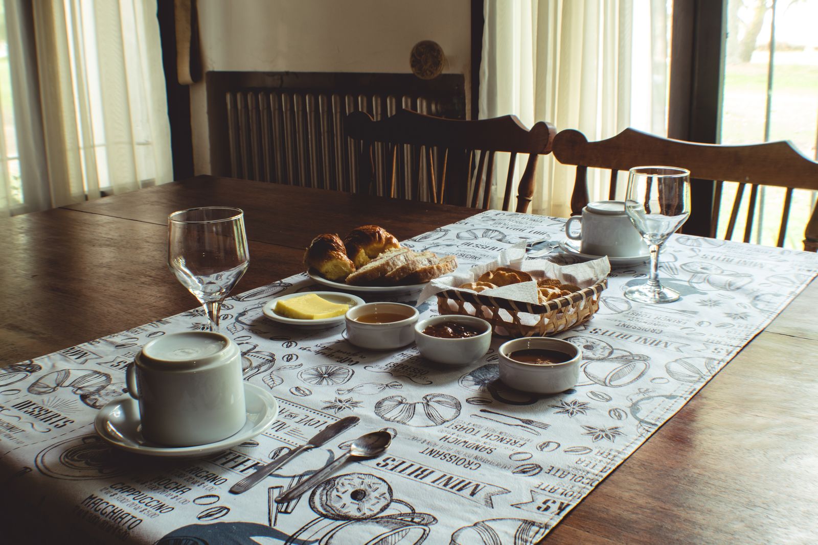 A breakfast table laid with bread, butter, jam and a teapot in a dining room