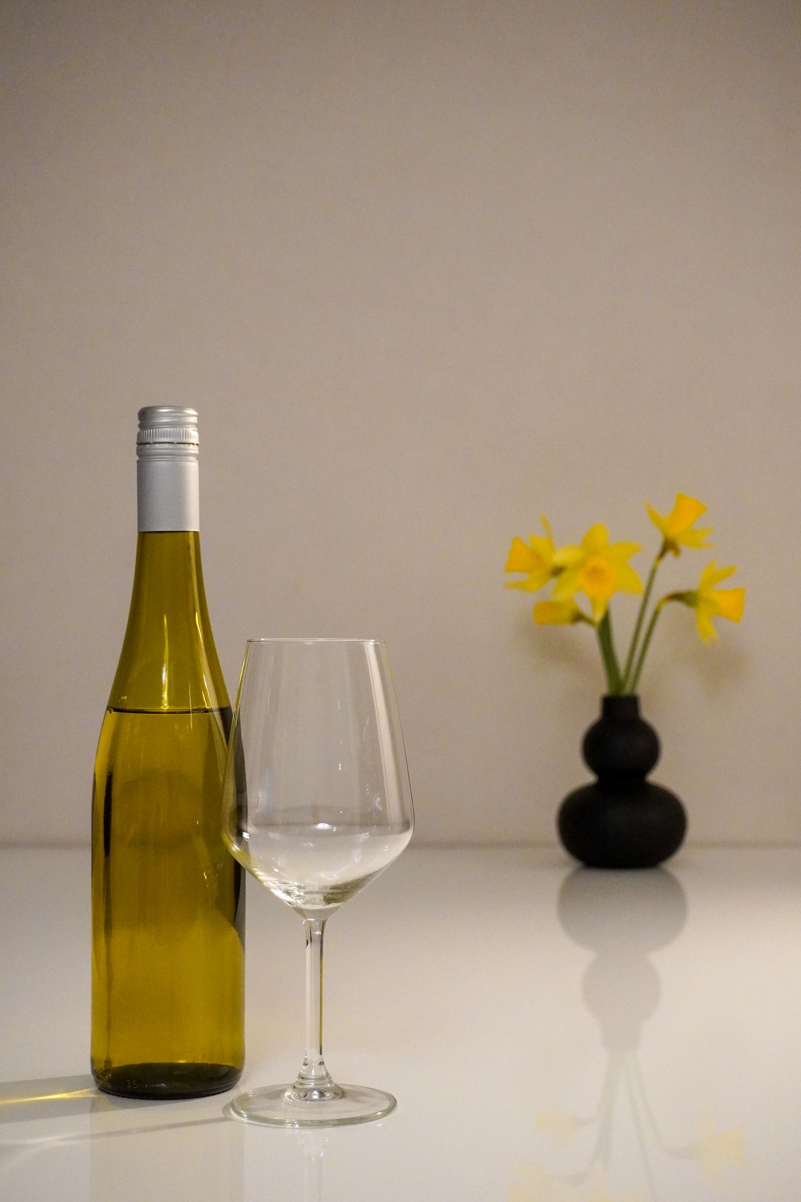 A bottle of white wine, an empty glass and a vase of yellow daffodils on a pale table