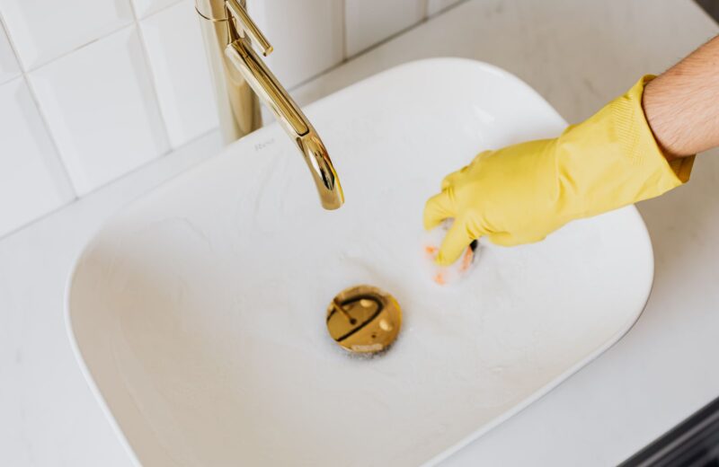 A hand in a yellow rubber glove cleaning a white basin with a sponge