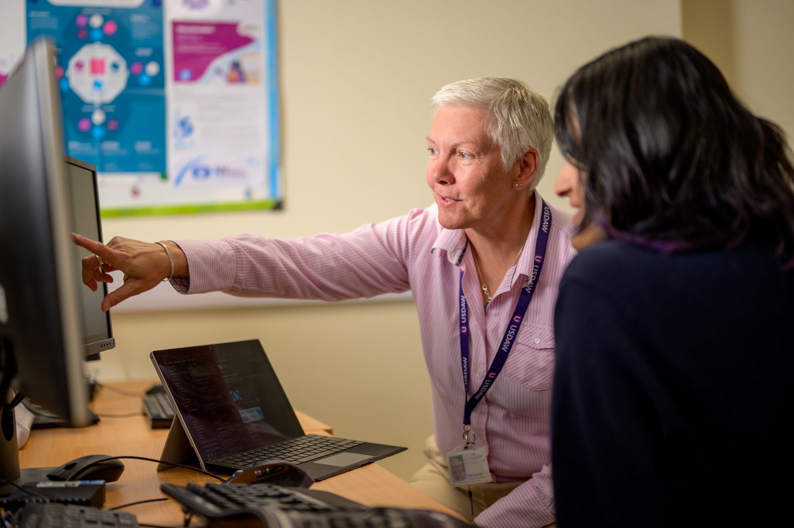 An IT engineer pointing at a monitor while a colleague follows on a laptop