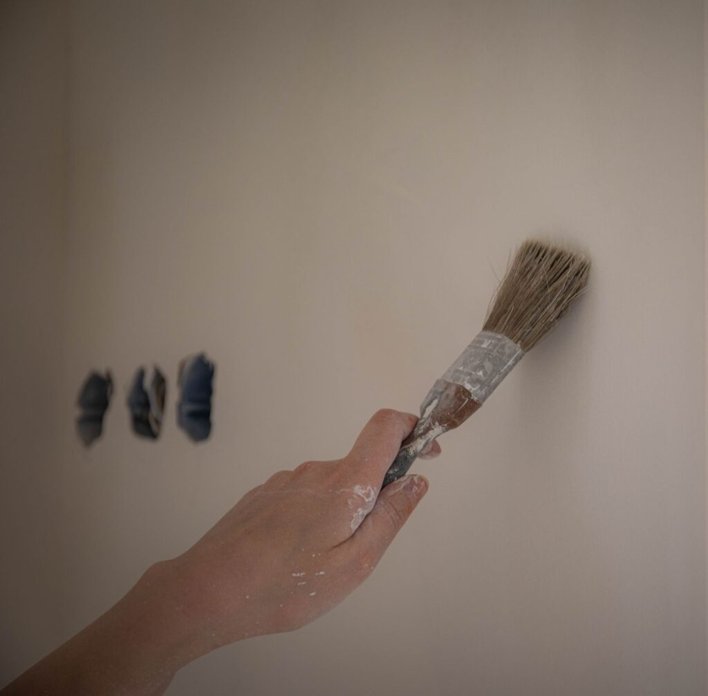 A hand painting a white wall beside three test patches of colour