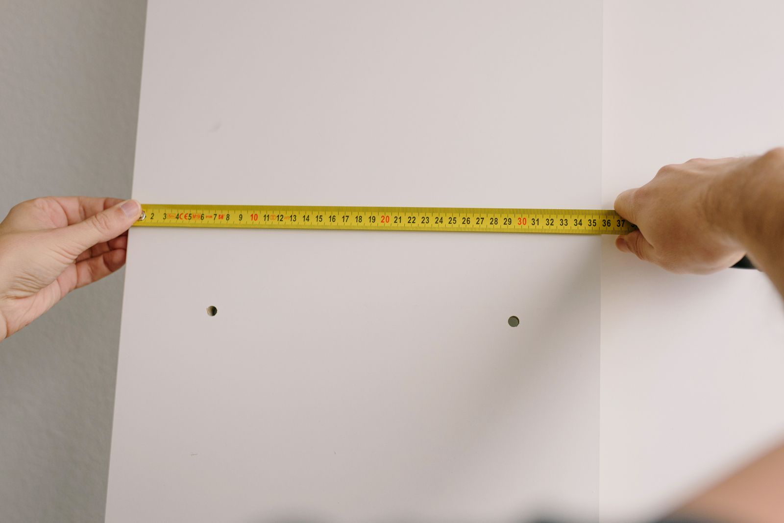 Two hands stretching a yellow tape measure across a white wall