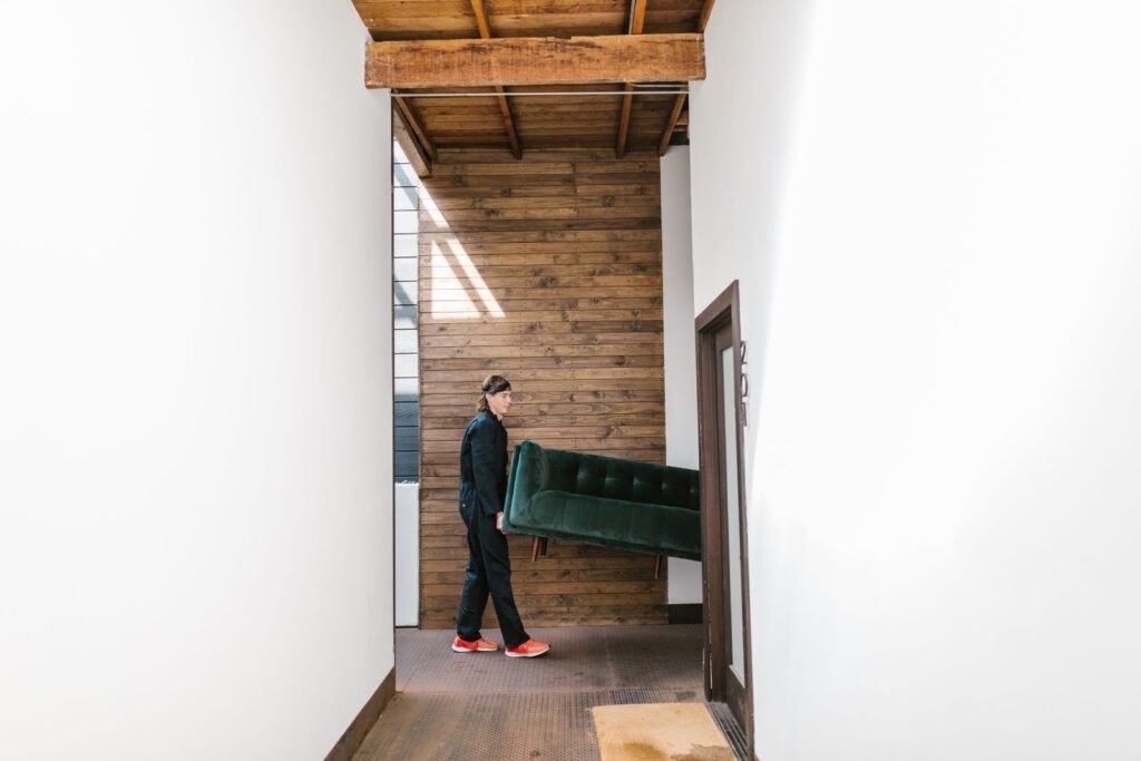 A delivery man carrying a green sofa along a hallway with wooden walls