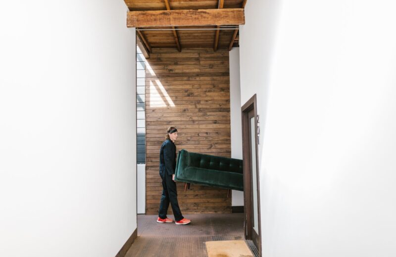 A delivery man carrying a green sofa along a hallway with wooden walls