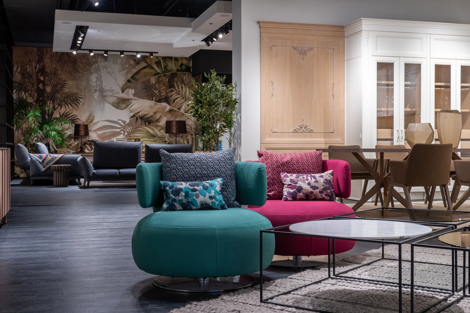 A furniture showroom with a teal velvet armchair, a pink sofa and low tables on a patterned rug