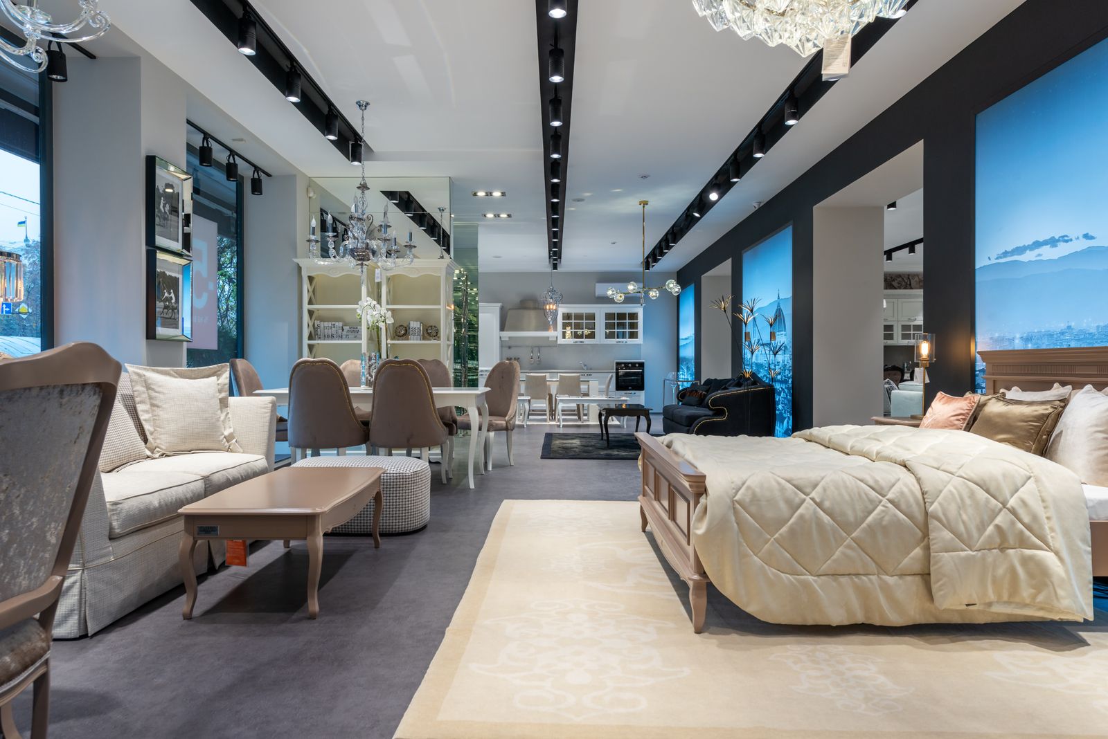 A furniture shop floor with a made bed, a soft grey sofa and a dining table under track lights