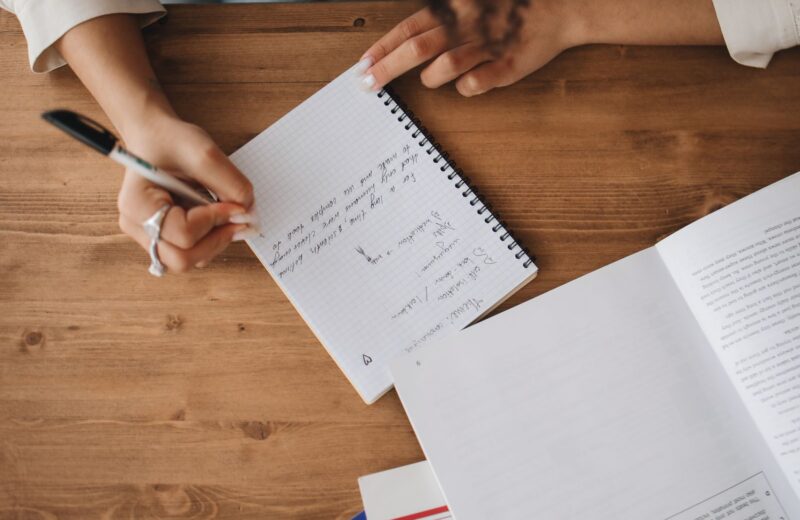 Hands taking notes in a notebook beside an open book on a wooden desk