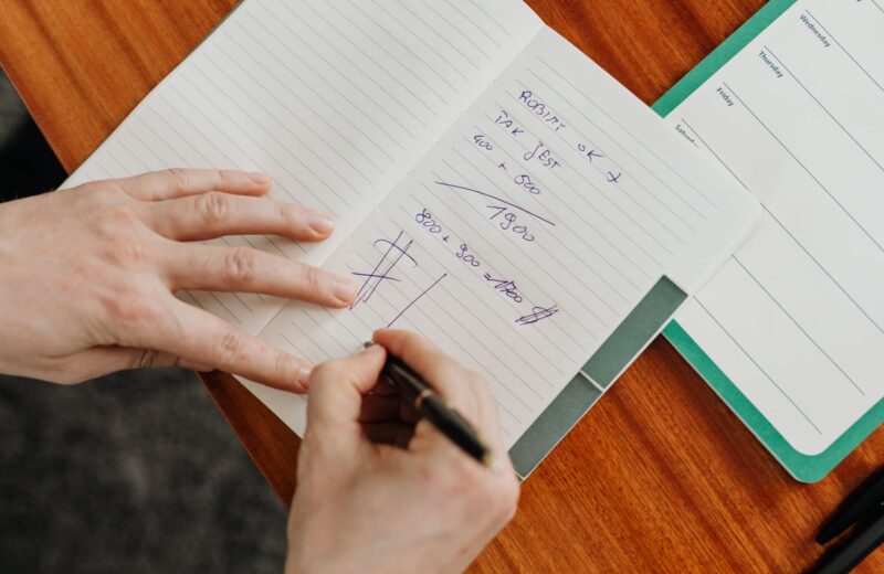 A hand writing sums in a notebook on a wooden desk