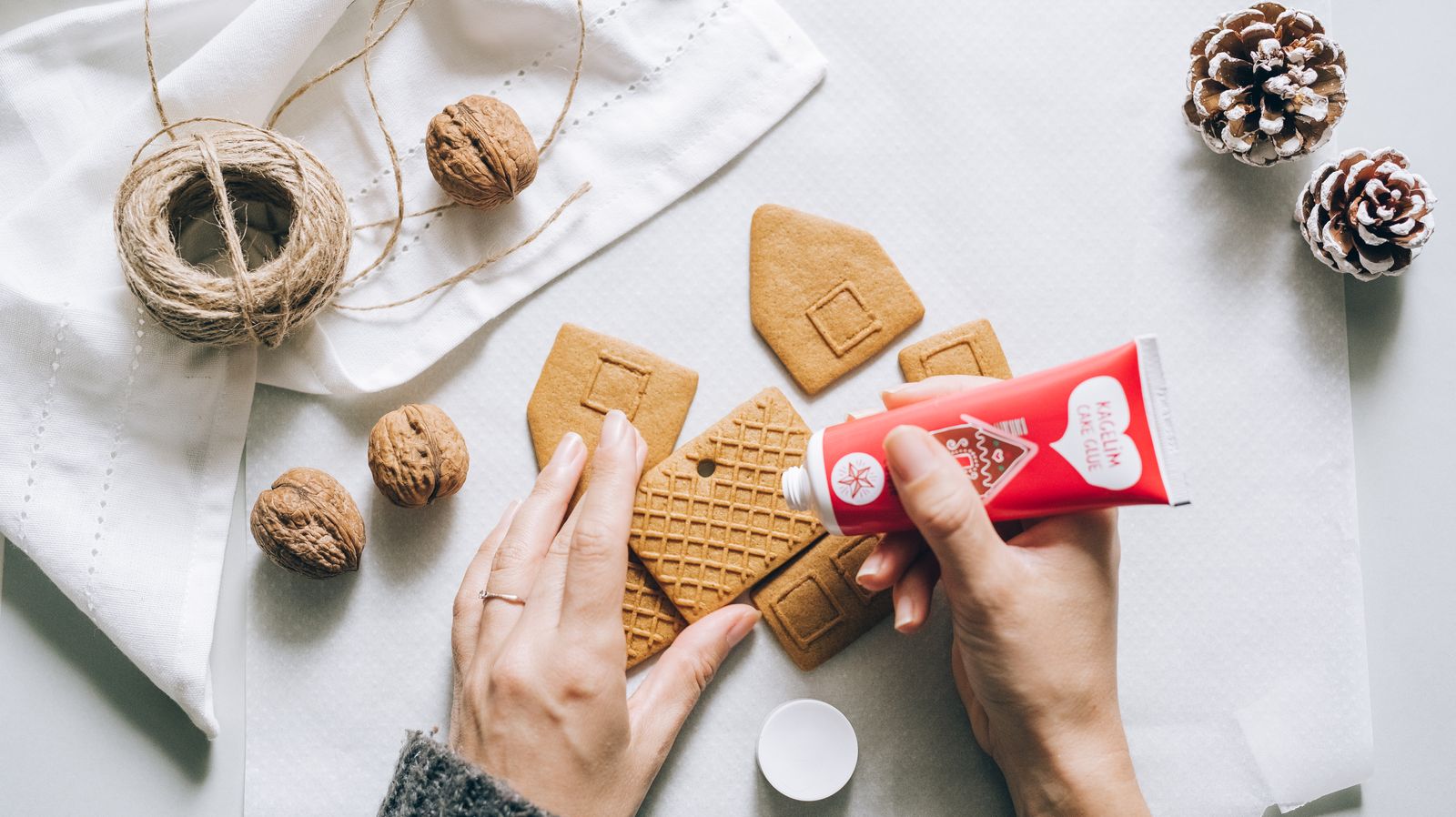 Hands piping red icing on gingerbread pieces on a white table