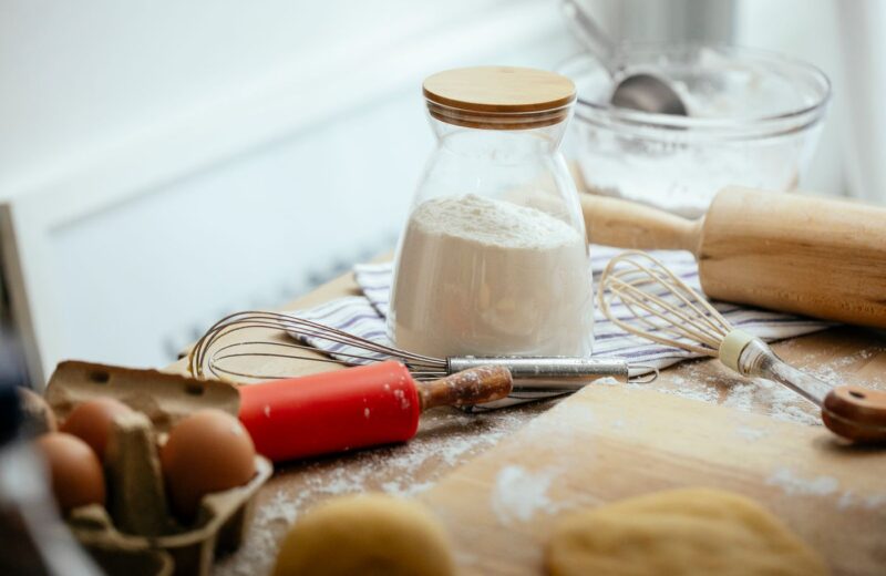 A rolling pin, a whisk, a jar of flour and eggs on a floured wooden table