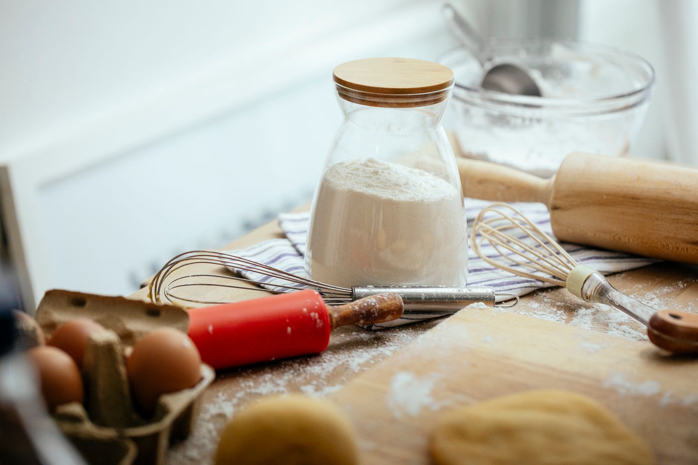 A rolling pin, a whisk, a jar of flour and eggs on a floured wooden table