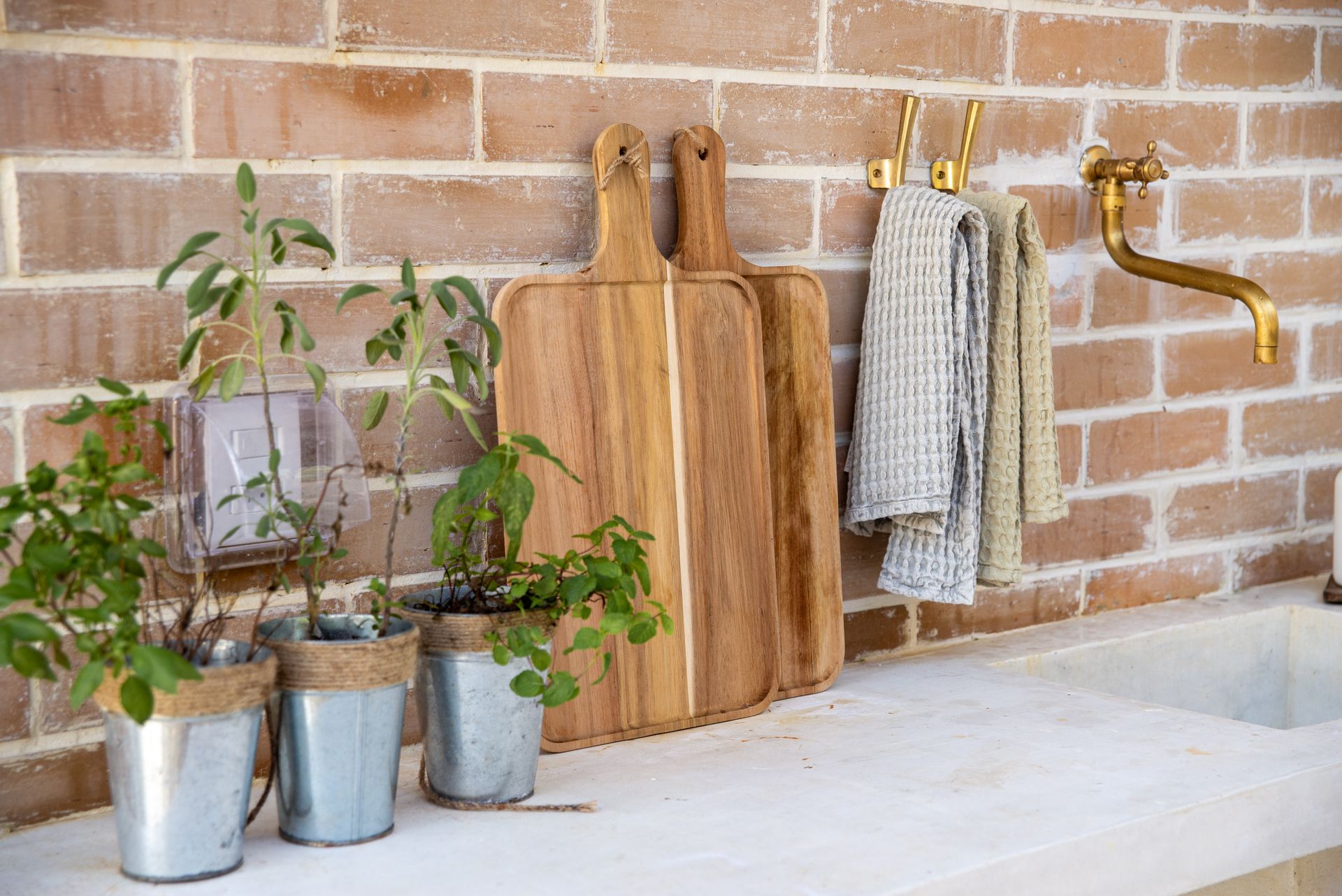 A brick kitchen wall with wooden cutting boards, a linen towel and pots of herbs on a white counter
