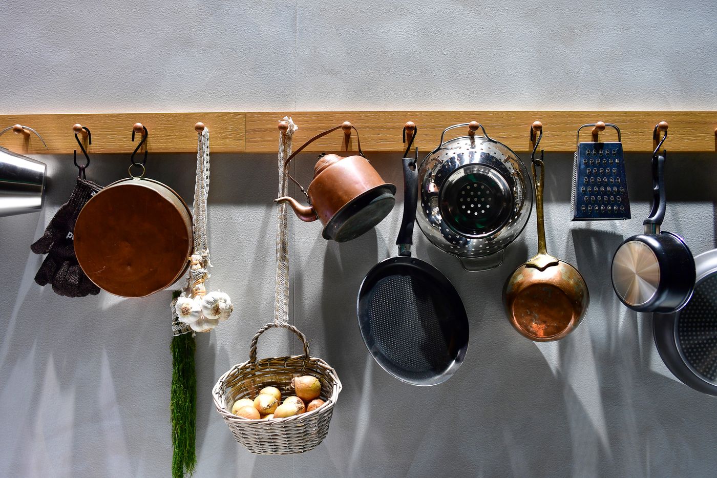 Copper and steel pans, a sieve and utensils hanging from a wooden rail on a grey wall