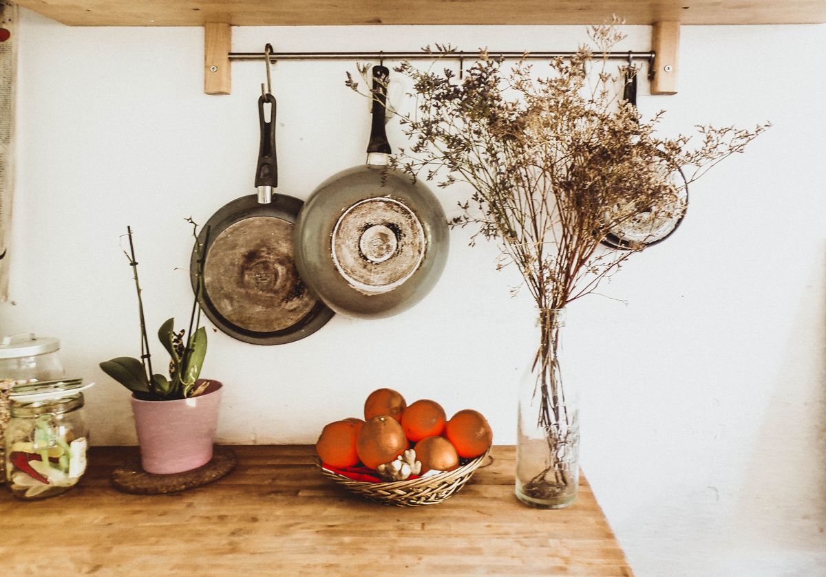 Two frying pans hanging on a white wall above a wooden counter with dried flowers and oranges