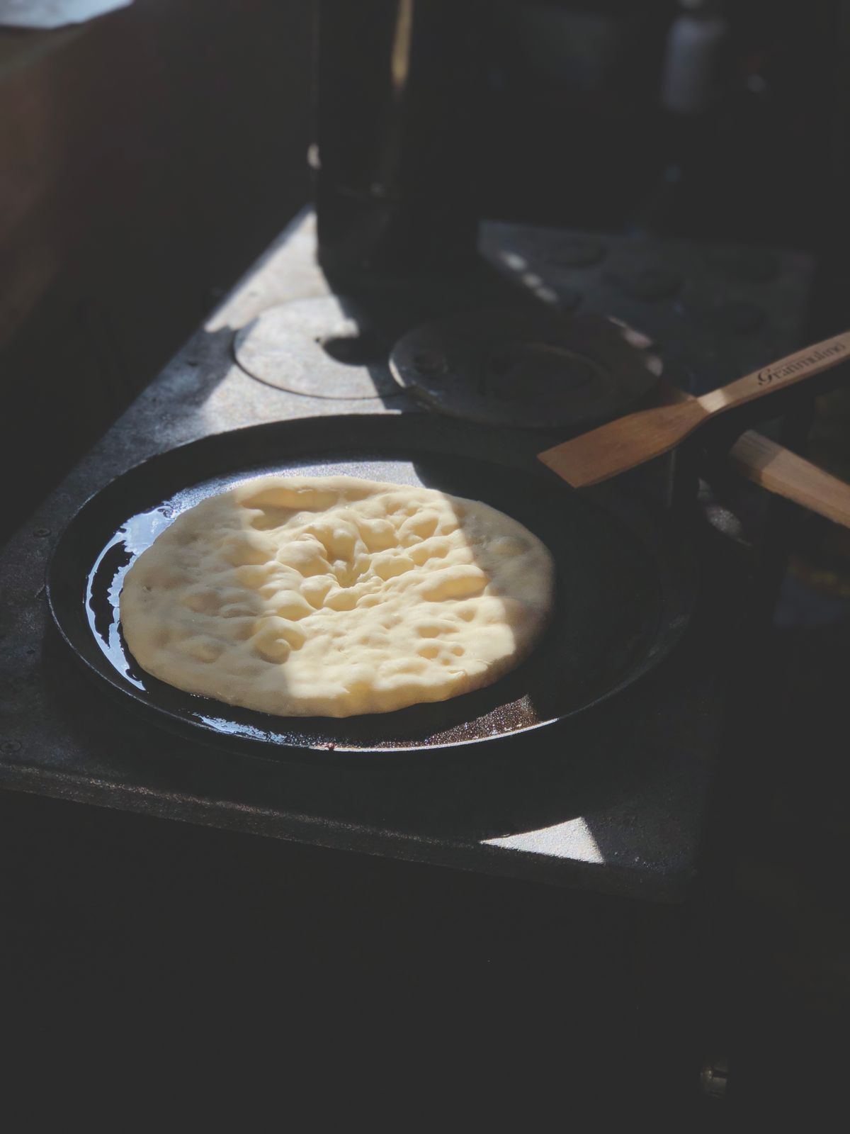 A flatbread cooking in a black cast iron skillet, a wooden spatula beside it