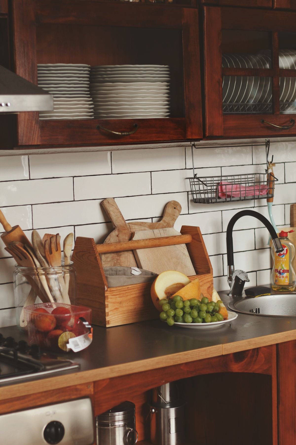 Wooden spoons and spatulas in a crate on a tiled kitchen counter with grapes and a sink