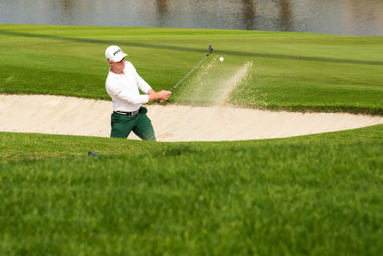 A golfer splashing a ball out of a greenside bunker beside a pond