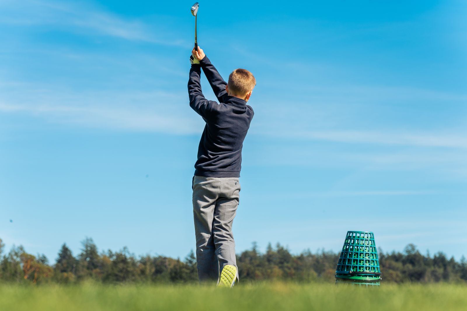 A young golfer seen from behind, finishing a full swing on a range under a blue sky