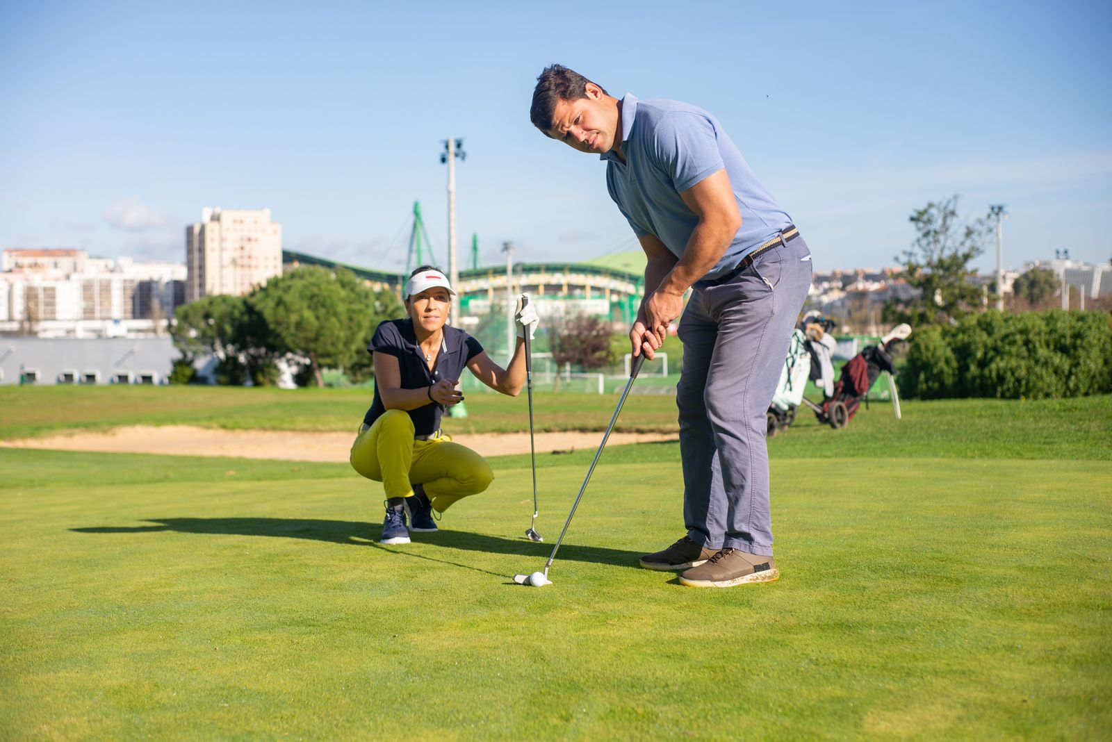 A coach crouches on a practice green and watches a pupil line up a short putt