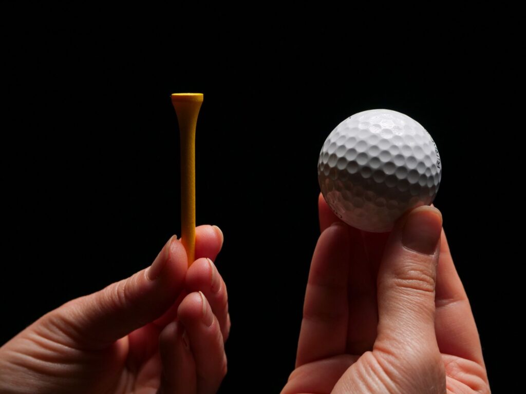 Two hands holding a wooden tee and a golf ball against a dark background